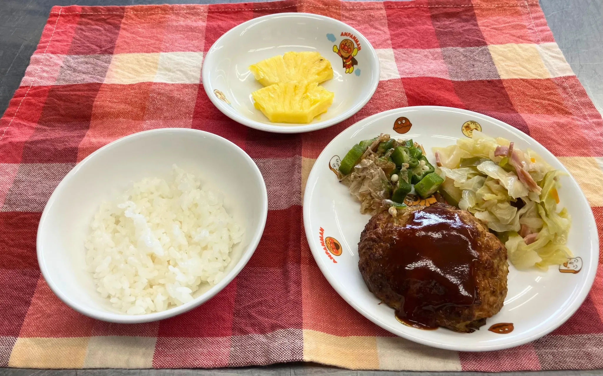 ある日の給食。ごはん、ハンバーグ、キャベツとベーコンの炒めもの、オクラのおかか和え、パイナップル